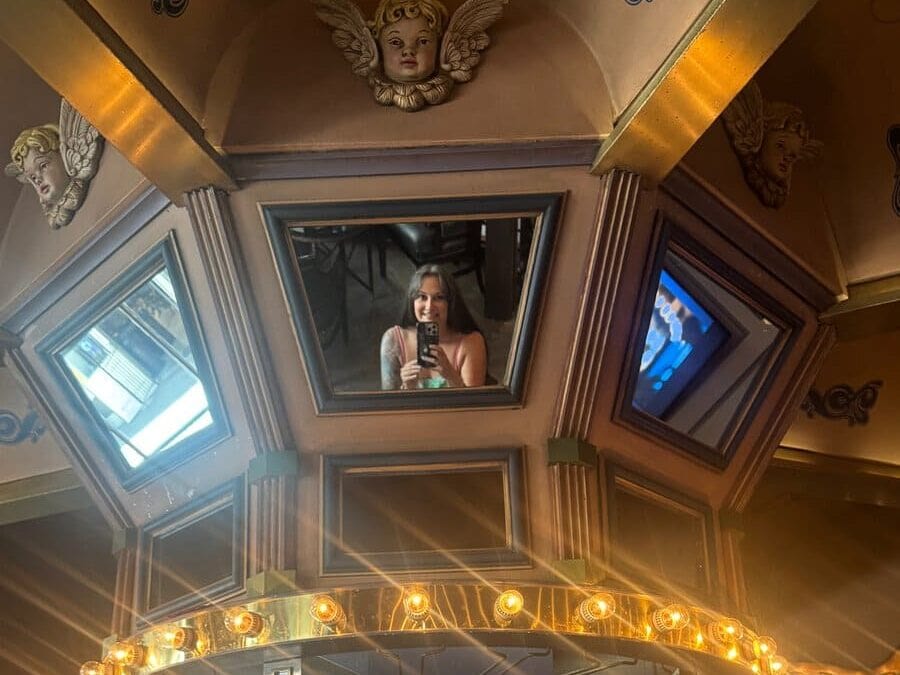 A person takes a photo of themselves in a mirrored ceiling above a bar, with various bottles and glasses displayed below and warm lights surrounding the structure—capturing one of the unique things to do alone in New Orleans for those seeking memorable solo adventures.