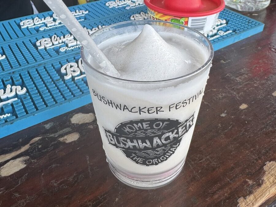 A frozen Bushwacker cocktail in a branded glass sits on a wooden bar with a blue mat and drink mixers in the background.