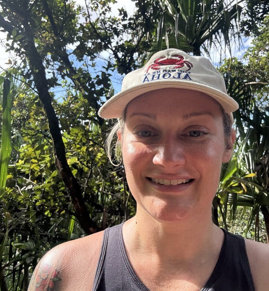 A person wearing a beige cap and black tank top smiles outdoors, surrounded by green trees and sunlight filtering through the leaves, capturing the vibrant energy of the Hanakapiai Falls Hike.