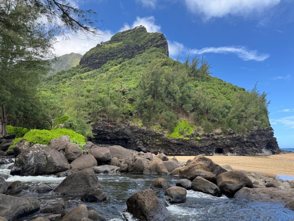 A lush green mountain rises above a sandy beach with dark rocks and a clear stream in the foreground, reminiscent of the landscapes seen along the Hanakapiai Falls Hike, all set under a bright blue sky with scattered clouds.