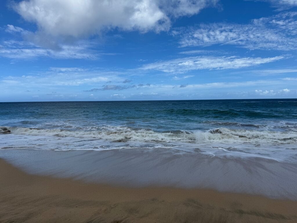 Waves gently crash onto a sandy beach under a partly cloudy blue sky, with the horizon line separating the deep blue ocean from the sky above—a tranquil scene reminiscent of the breathtaking vistas experienced along the Hanakapiai Falls Hike.