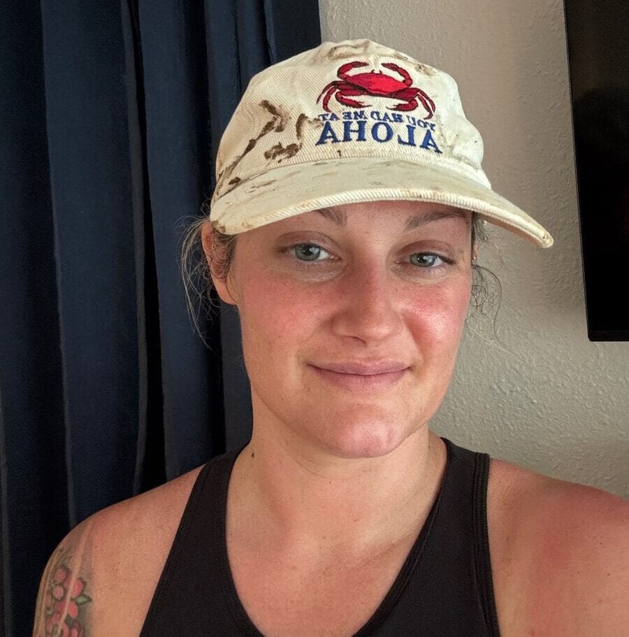 A person wearing a white hat with a red crab and IM CRABBY ALOHA text, black tank top, and a visible floral tattoo on their shoulder, smiling in front of a cream wall and dark blue curtains—capturing the laid-back spirit after completing the Hanakapiai Falls Hike.