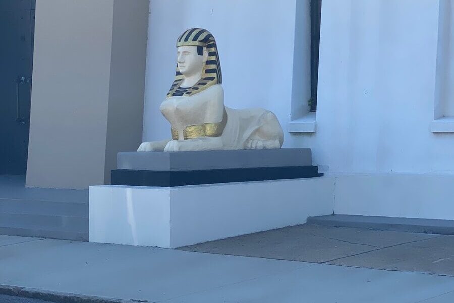 A small sphinx statue with a gold and black striped headdress sits on a white platform outside a light-colored building—an unexpected find for those enjoying solo travel along Alabama's Gulf Coast.
