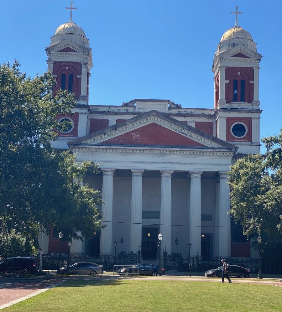 A large red-brick church with two bell towers topped by gold domes and crosses, white columns at the entrance, and trees partially framing the building on a sunny day—a captivating sight for solo travel along Alabama's Gulf Coast.