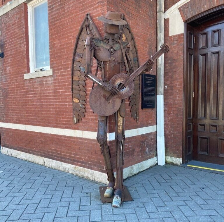 A metal sculpture of an angel with wings, wearing a hat and playing a guitar, stands on a brick sidewalk by a red brick building with a wooden door—an inspiring find for solo travel along Alabama's Gulf Coast.