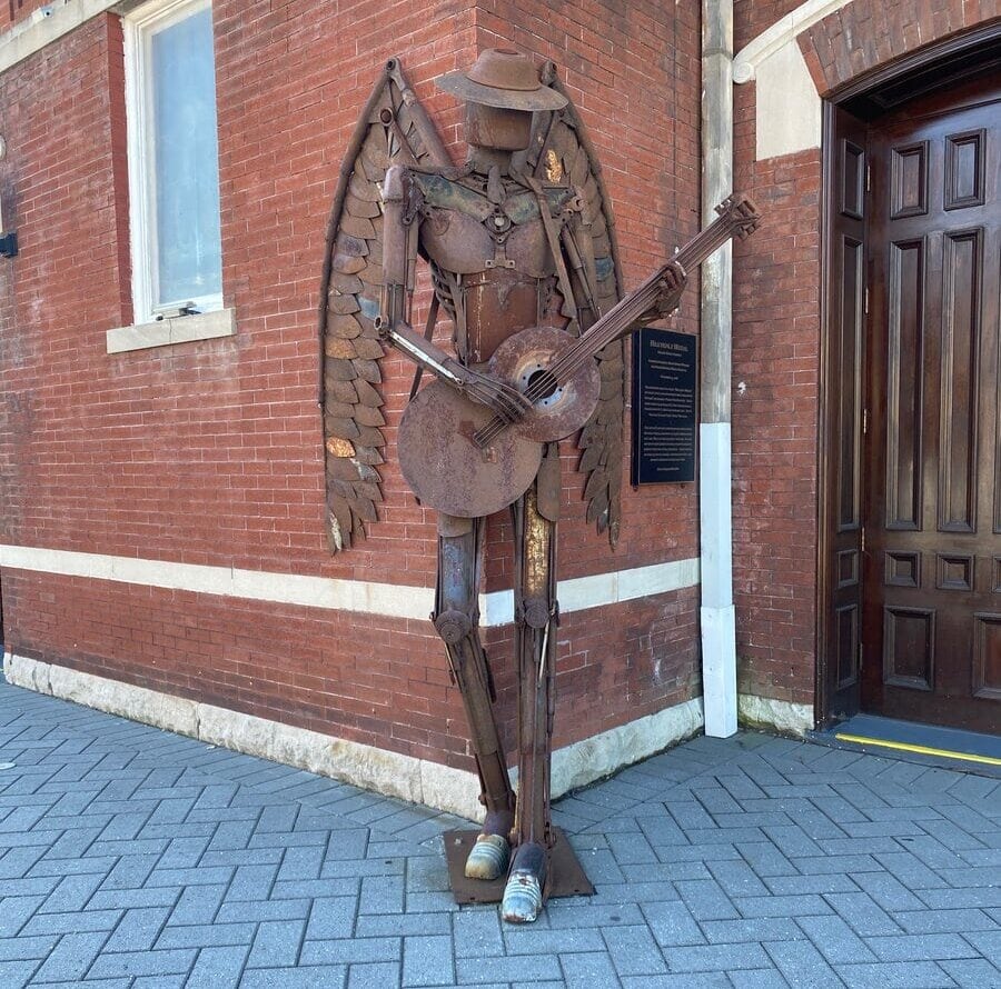 A metal sculpture of an angel with wings, wearing a hat and playing a guitar, stands on a brick sidewalk by a red brick building with a wooden door—an inspiring find for solo travel along Alabama's Gulf Coast.